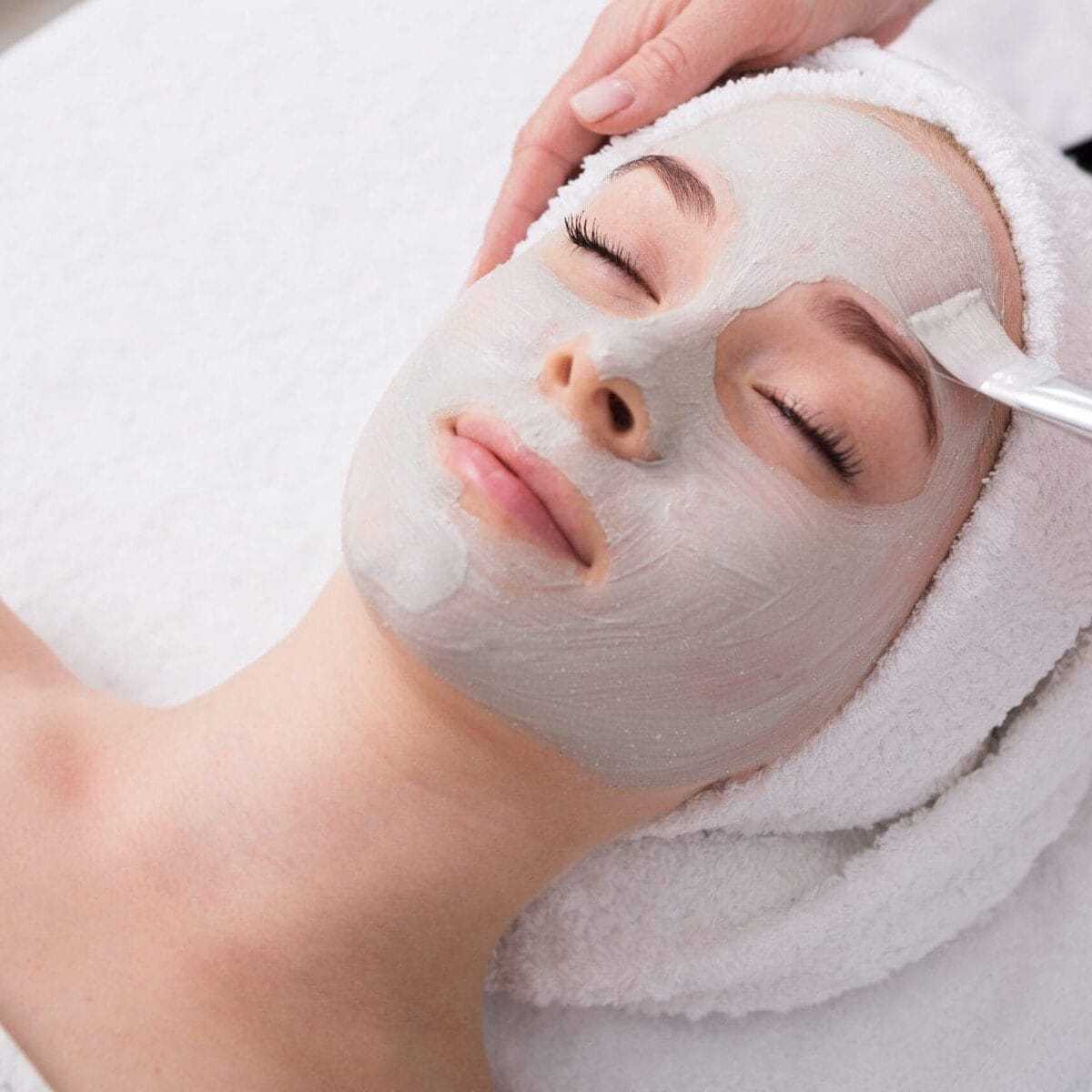 Woman receiving a relaxing facial treatment with a clay mask applied by a cosmetologist.
